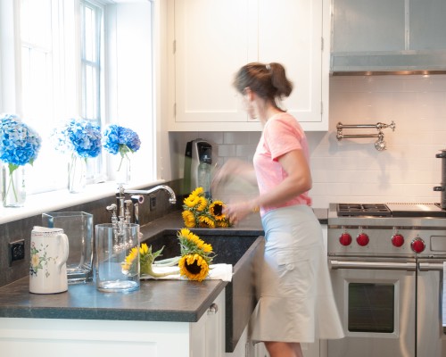 A woman in the kitchen with flowers on the counter.