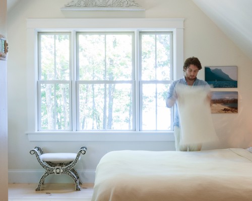 A man is holding pillows in the corner of a bedroom.